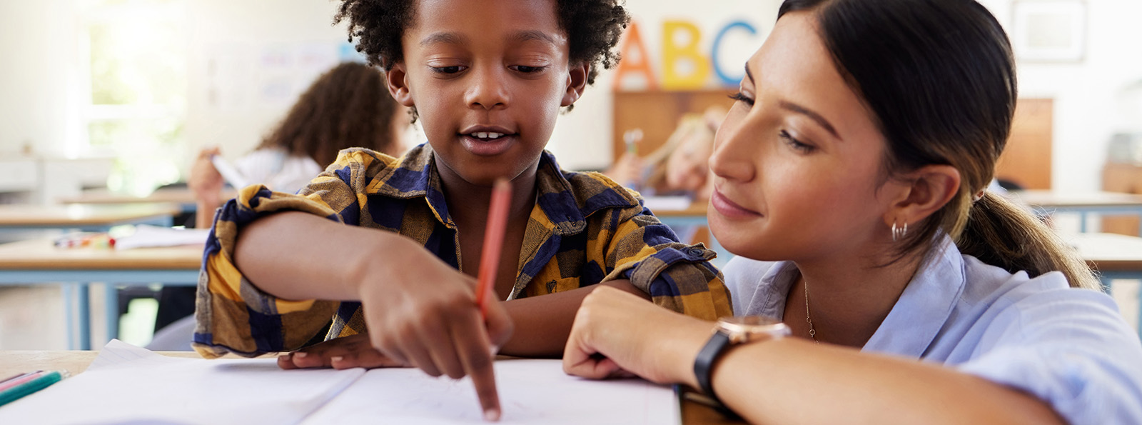 a teacher helps a young student while he holds a pencil and points at his paper