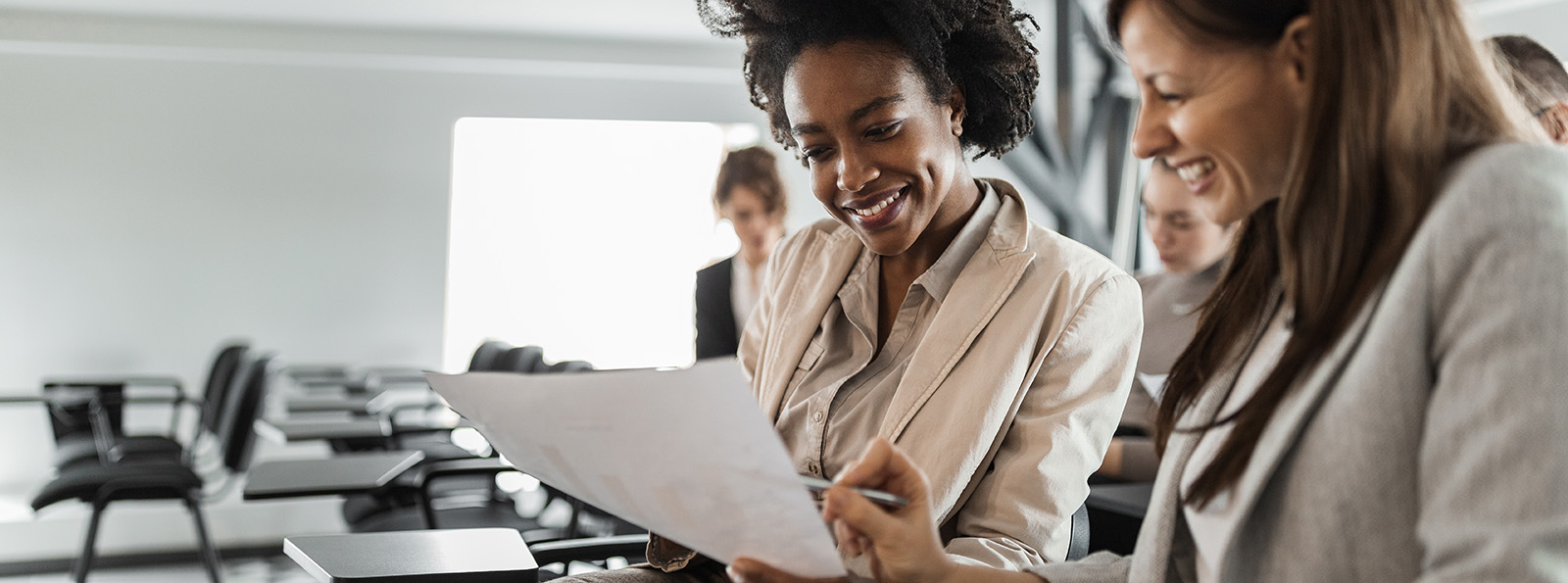 two professional women smiling as they look at the same sheet of paper in an office