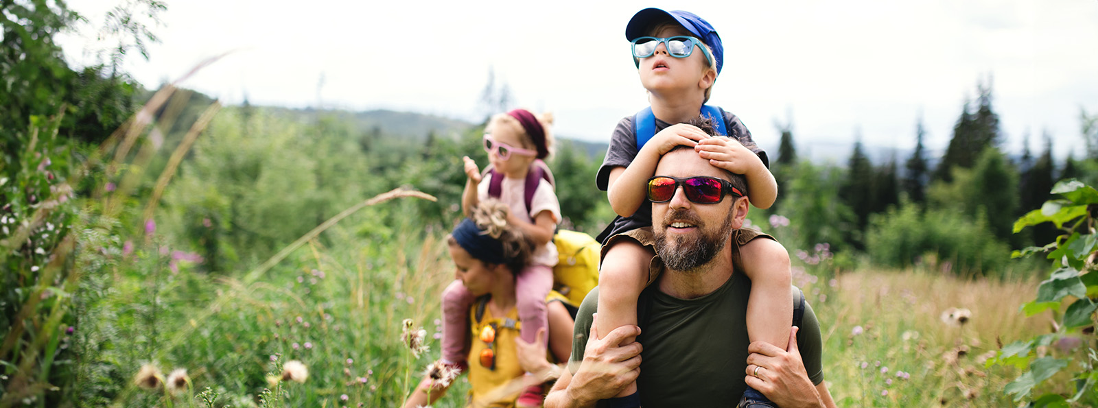 a parents with young kids on their shoulders hikes through a grassy area 