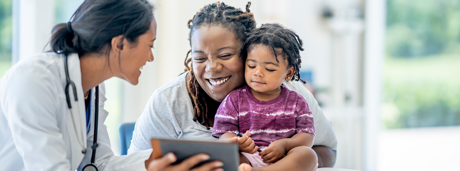 a doctor with a stethoscope holds a digital tablet for a smiling mother and her daughter to look at