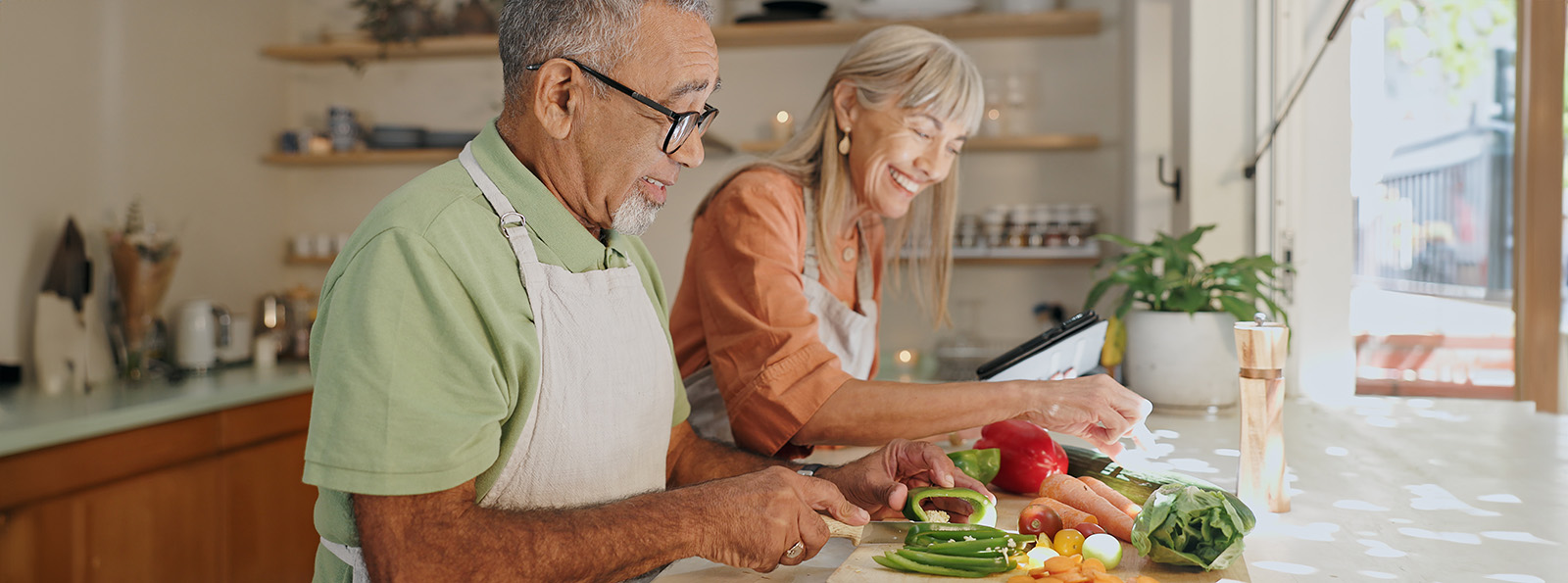 an older couple chopping vegetables at a counter near a window