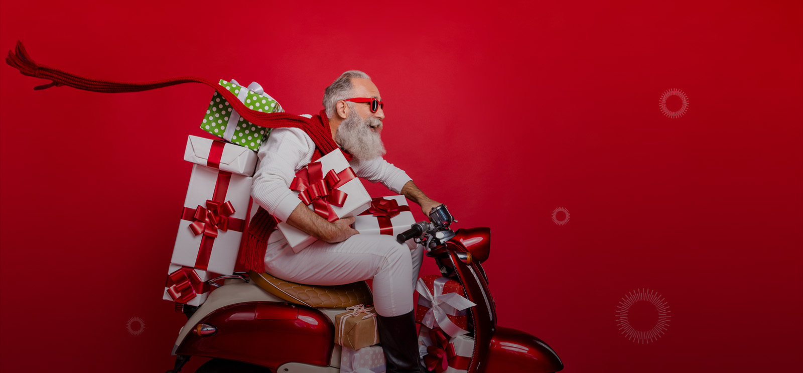A man that looks like Santa Claus riding a red scooter, holding several wrapped gifts and smiling while wearing sunglasses