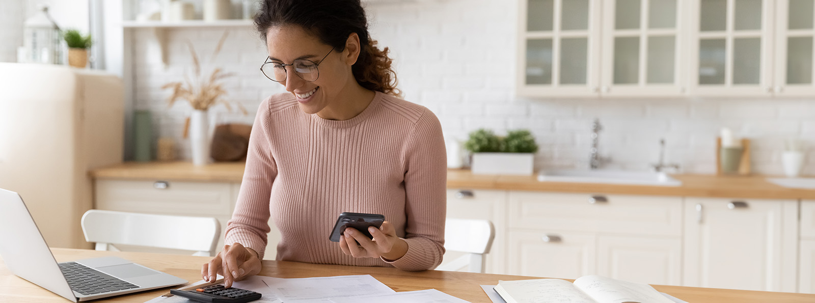 A woman using a calculator, and a phone while next to a laptop with pieces of paper and books open on the kitchen table