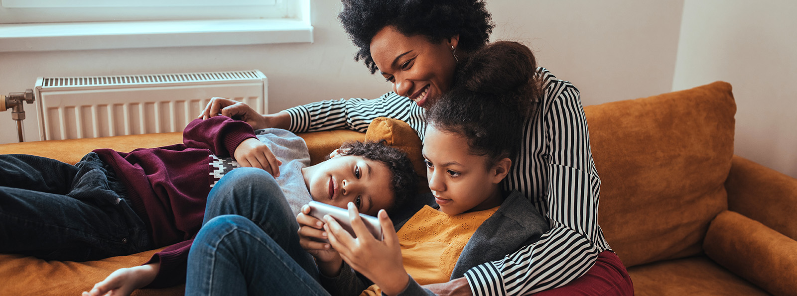 a smiling mother on a bed with her two kids as one of them is using a mobile phone