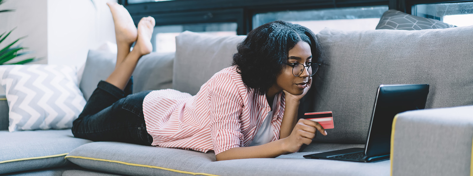 a woman lays on her couch with her credit card in hand while shopping on her laptop